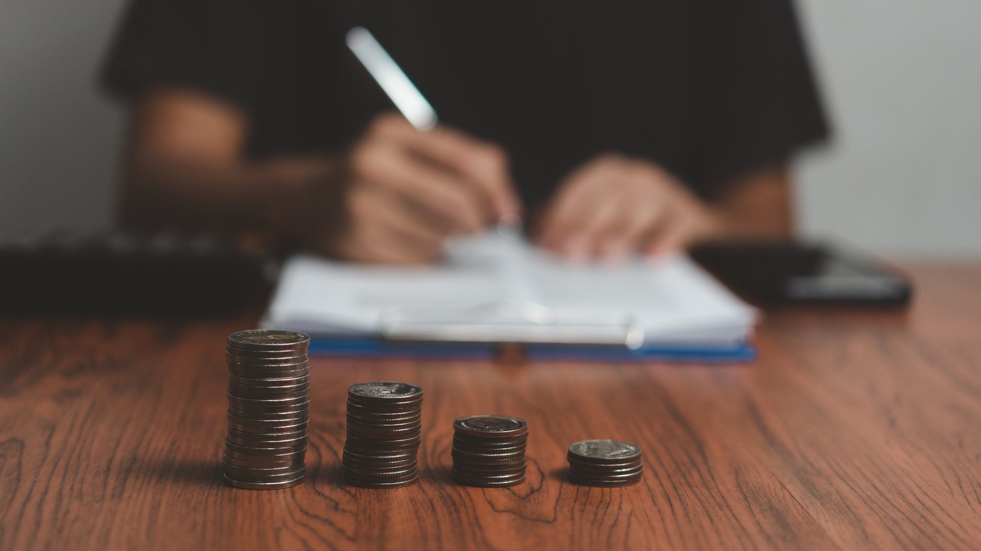 Pilas de monedas decrecientes en una mesa de madera; al fondo, alguien rellena documentos – sueldo del E-commerce Manager