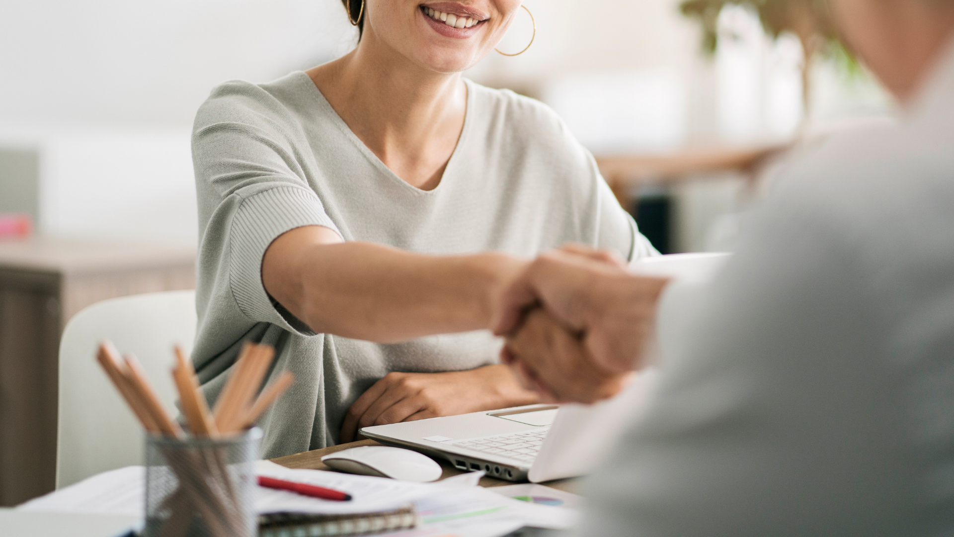Mujer sonriente da la mano a su interlocutor ante un escritorio con portátil – contratación de un especialista en e-commerce